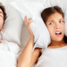 Annoyed woman in bed, pressing a pillow to her ears, lying next to a snoring man