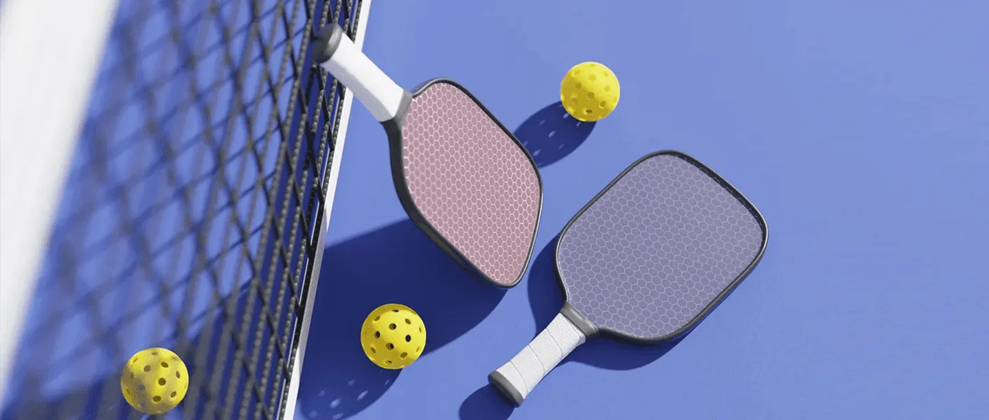 Two pickleball paddles and three yellow perforated balls are on a blue court near a net