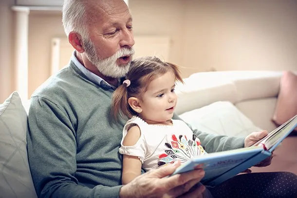 Elderly man with a beard reads a picture book to a young girl with plaits while sitting on a sofa.